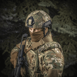 Sotilas pukeutuneena M‑Tac Hjelmcover Shroud Elite MULTICAMissa TOR-D-kypärälle, ase mukanaan metsämaisemassa.