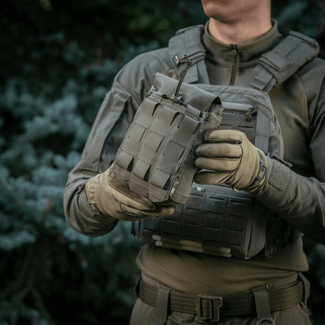 M‑Tac Medic Pouch Vertical Large Elite käytössä, pidettynä sotilasasuisen henkilön toimesta taktisen taustan edessä.