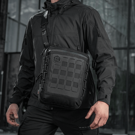Mies mustassa takissa kantaa M-Tac Tablet Bag Laser Cut Hex -laukkua, tukeva ja käytännöllinen EDC- ja piilotuskantolaukku.