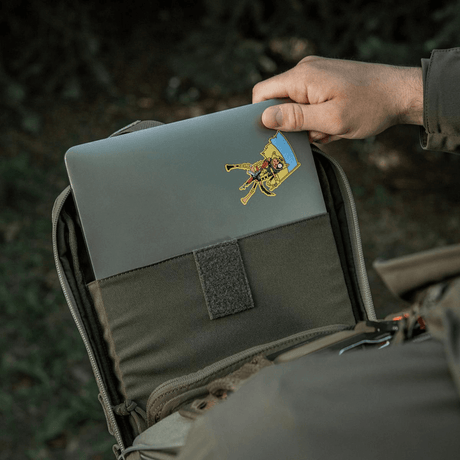 Henkilö laittaa läppäriä M-Tac Tablet Bag Laser Cut Hex -laukkuun, Laser Cut MOLLE, Cordura-materiaali tabletille ja piilotetulle aseelle.