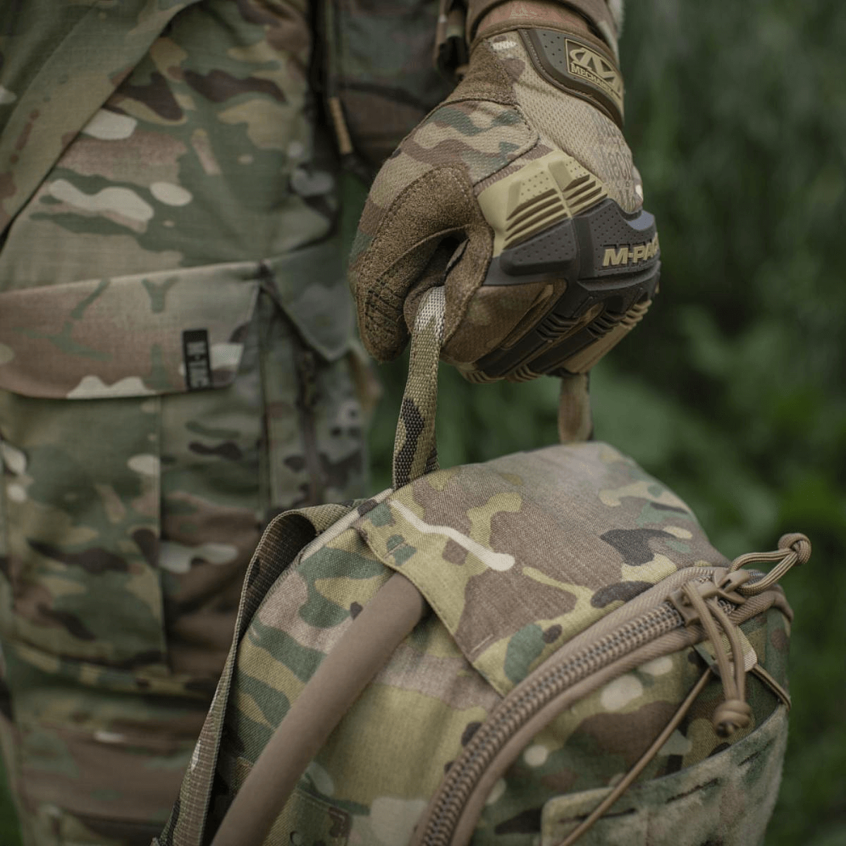M-Tac STURM Elite -reppu camo-muotoilulla ja MOLLE-järjestelmällä, pidetty henkilön toimesta armeijavaatteissa ja taktisisissa käsineissä, 15 l tilavuus.