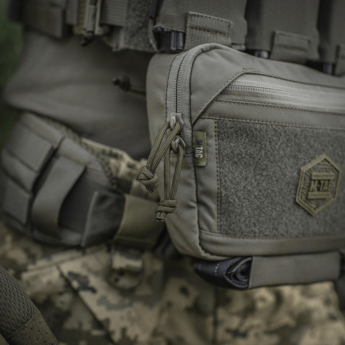 M-Tac Large Elite Gen.II olkalaukku Cordura 500D:stä YKK-vetoketjuilla, sopii taktiseen käyttöön, nähty maastotaustalla.