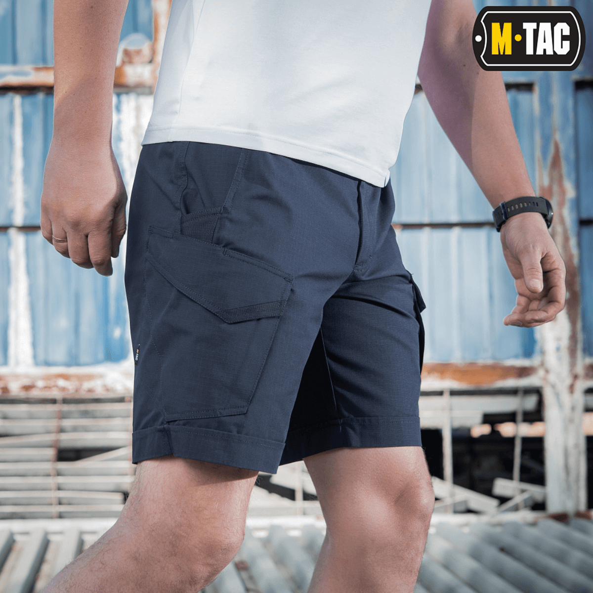 Mies pukeutuneena M-Tac Shorts Aggressor Shortiin sinisenä, sopii taktiseen käyttöön lämpiminä päivinä, valmistettu joustavasta polycotton rip-stop -materiaalista.