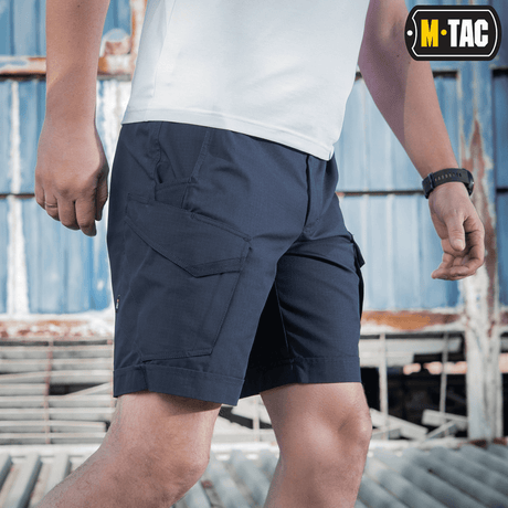 Mies pukeutuneena M-Tac Shorts Aggressor Shortiin sinisenä, sopii taktiseen käyttöön lämpiminä päivinä, valmistettu joustavasta polycotton rip-stop -materiaalista.