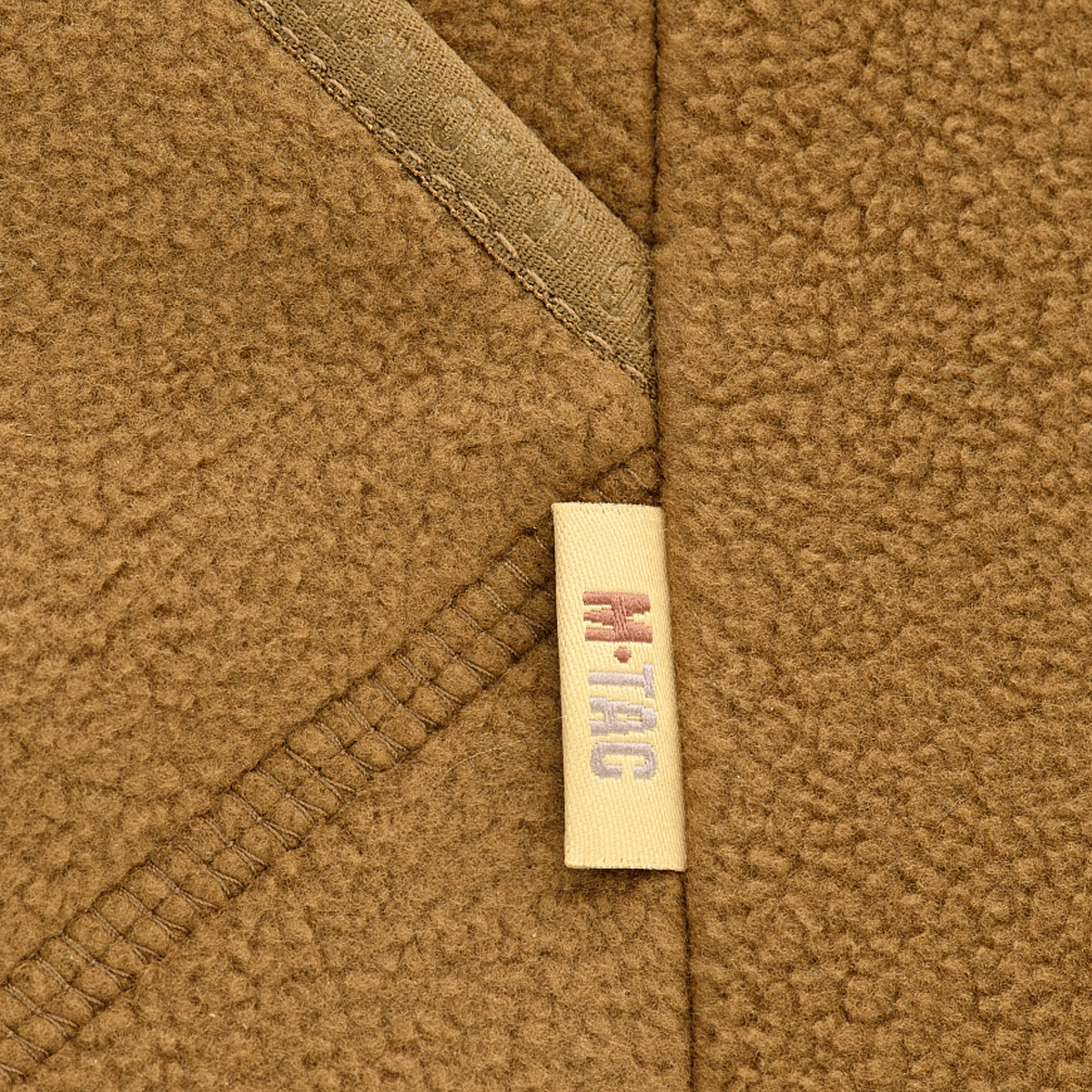 Lähikuva M-Tac Lite Microfleece Hættetrøje -tuotteesta ruskeassa mikrofleecessä, M-Tac-logo sauman kohdalla, fokus materiaalissa ja yksityiskohdissa.