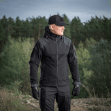Mies M-Tac Norman Windblock -fleecetakissa metsäalueella, sopii kylmään ja tuuliseen säähän, näyttää takin toiminnallisuuden ja muotoilun.
