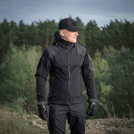 Mies M-Tac Norman Windblock -fleecetakissa metsäalueella, sopii kylmään ja tuuliseen säähän, näyttää takin toiminnallisuuden ja muotoilun.