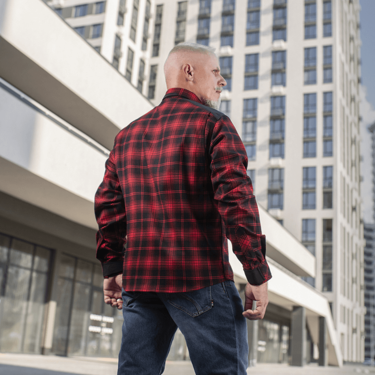 Mies pukeutuneena M-Tac Redneck -paitaan punaruutuisena, modernin rakennuksen edessä, sopii työhön ja vapaa-aikaan mukavalla istuvuudella.
