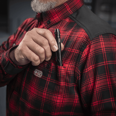 Mies pukeutuneena M-Tac Redneck -paitaan punaisessa ruudussa ja mustilla vahvikkeilla, pitää taskulamppua rintataskussa.