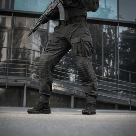 Mies käyttää M-Tac STURM NyCo Extreme Gen.II -housuja aseen kanssa modernin rakennuksen edessä.