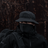 Henkilö pukeutuneena M-Tac Panama Rip-Stop army-hattuun mustana, puolijäykkä lippa ja tuuletusreiät, sovitettu maastokuviointiin ja tuulensuojaan.