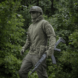 Henkilö pukeutuneena M-Tac Division Gen.II Windblock Fleecejakke, suunniteltu maksimaaliseen lämpöön ja tuulisuojaan, vaeltaa metsäalueella.