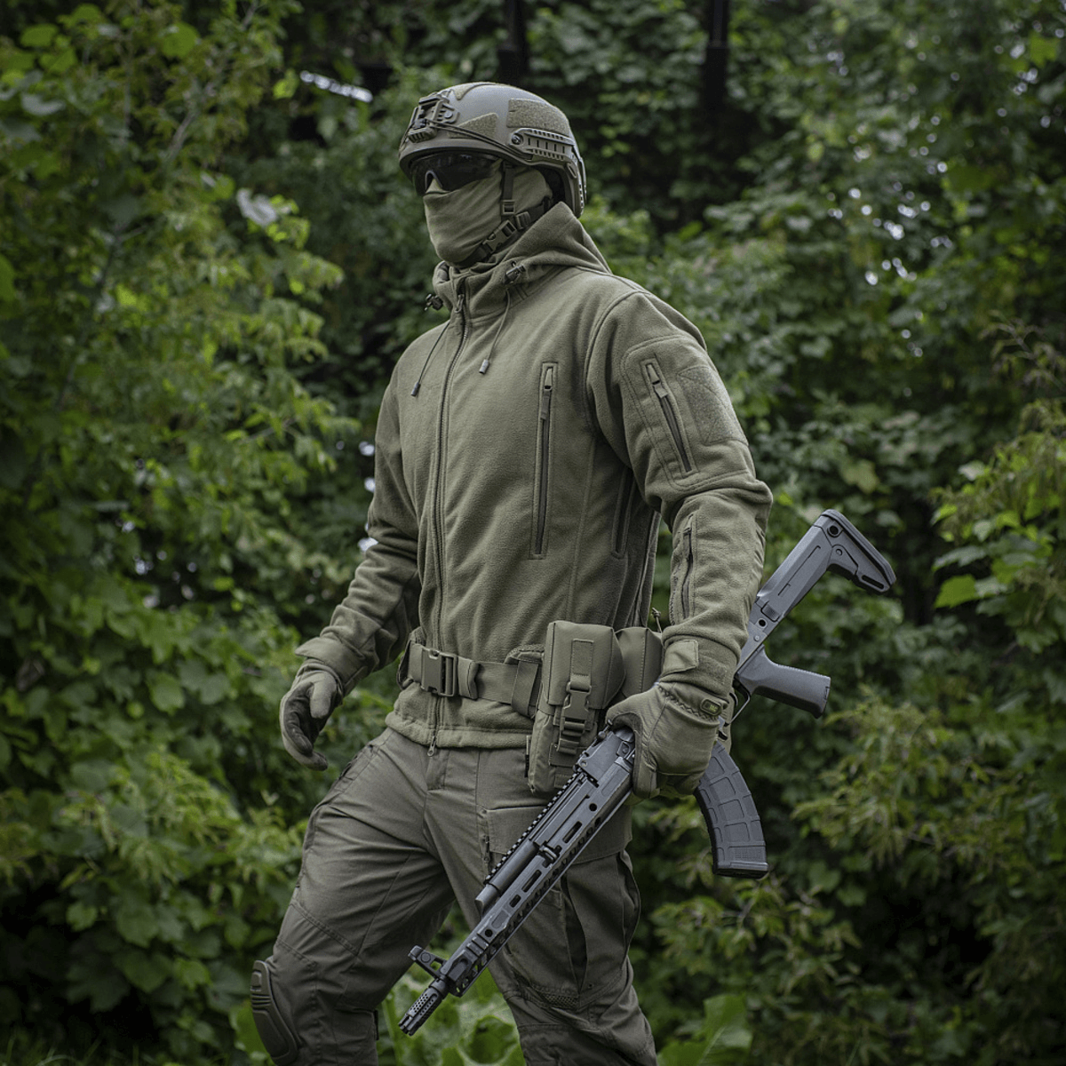 Henkilö pukeutuneena M-Tac Division Gen.II Windblock Fleecejakke, suunniteltu maksimaaliseen lämpöön ja tuulisuojaan, vaeltaa metsäalueella.