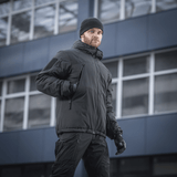 Mies pukeutuneena M-Tac Alpha Gen.III Pro talvitakkiin, musta, sopii lämpötiloihin jopa -20°C, modernin rakennuksen edessä.