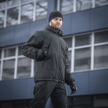 Mies pukeutuneena M-Tac Alpha Gen.III Pro talvitakkiin, musta, sopii lämpötiloihin jopa -20°C, modernin rakennuksen edessä.