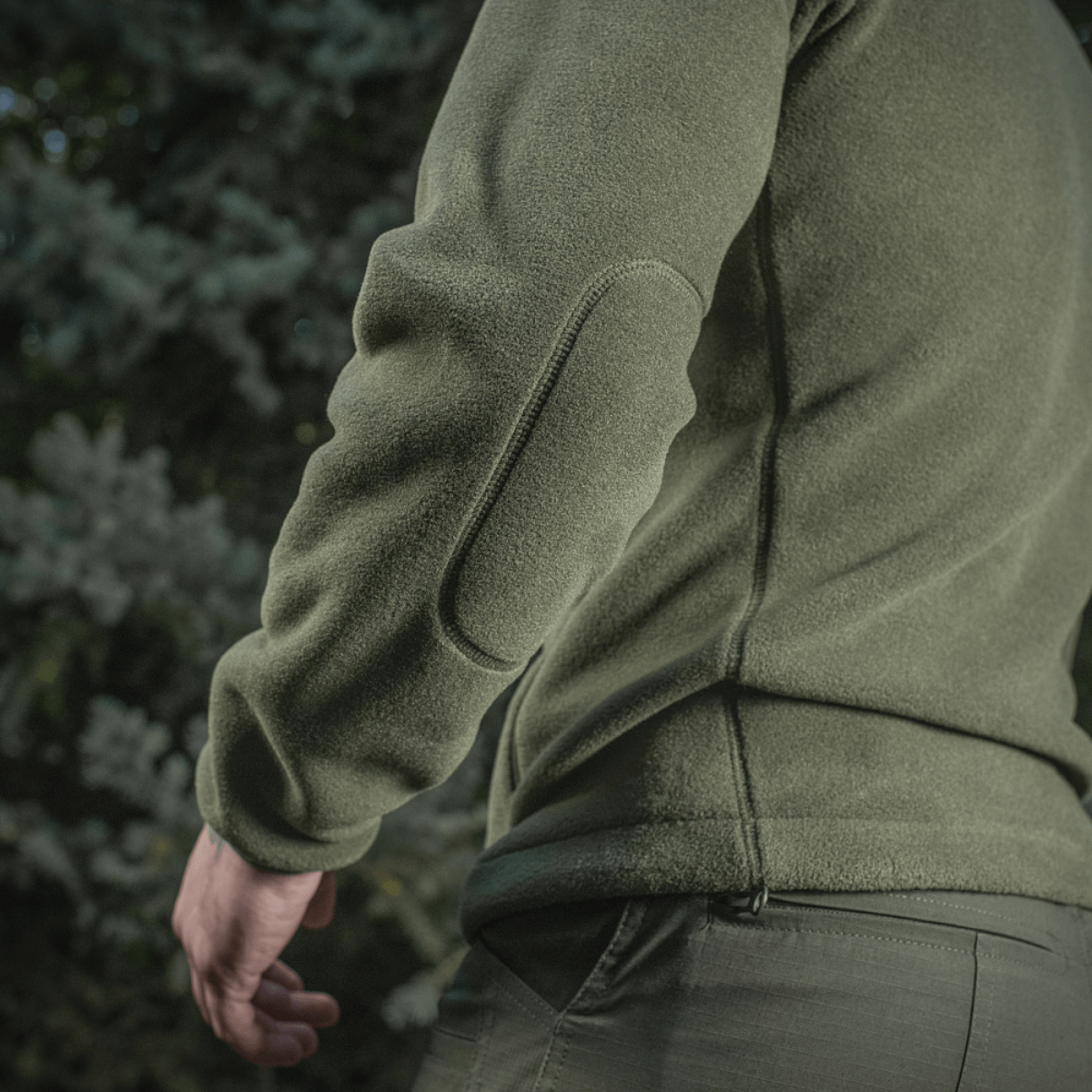 Mies pukeutuneena M-Tac Nord Polartec -fleecetakkiin vihreänä, suunniteltu tyyliin ja toimivuuteen, nähtynä takaa ulkoilmassa puiden ympäröimänä.