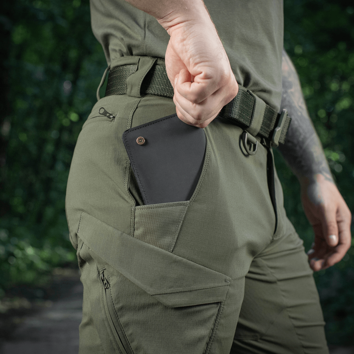 Mies pukeutuneena M-Tac Aggressor Summer Flex Shorts -shortseihin metsässä, käsi taskussa näyttää puhelimen säilytyksen. Tuoteavainsana: M-Tac Aggressor Summer Flex Shorts.