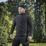 Mies pukeutuneena M-Tac Sprint Fleece Sweatshirt Polartec mustana rehevässä metsäympäristössä, korostaa tyylikästä toiminnallisuutta ja mukavuutta.