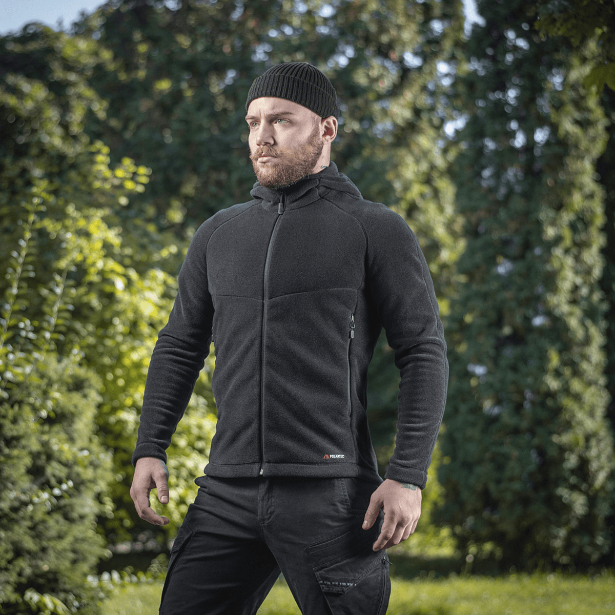 Mies pukeutuneena M-Tac Sprint Fleece Sweatshirt Polartec mustana rehevässä metsäympäristössä, korostaa tyylikästä toiminnallisuutta ja mukavuutta.