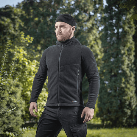 Mies pukeutuneena M-Tac Sprint Fleece Sweatshirt Polartec mustana rehevässä metsäympäristössä, korostaa tyylikästä toiminnallisuutta ja mukavuutta.