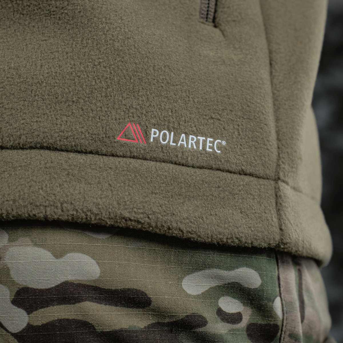 Yksityiskohta M-Tac Combat Fleece Polartec -takista, Polartec-logo maastokuvioisella taustalla, korostaa takkia korkealaatuisena.