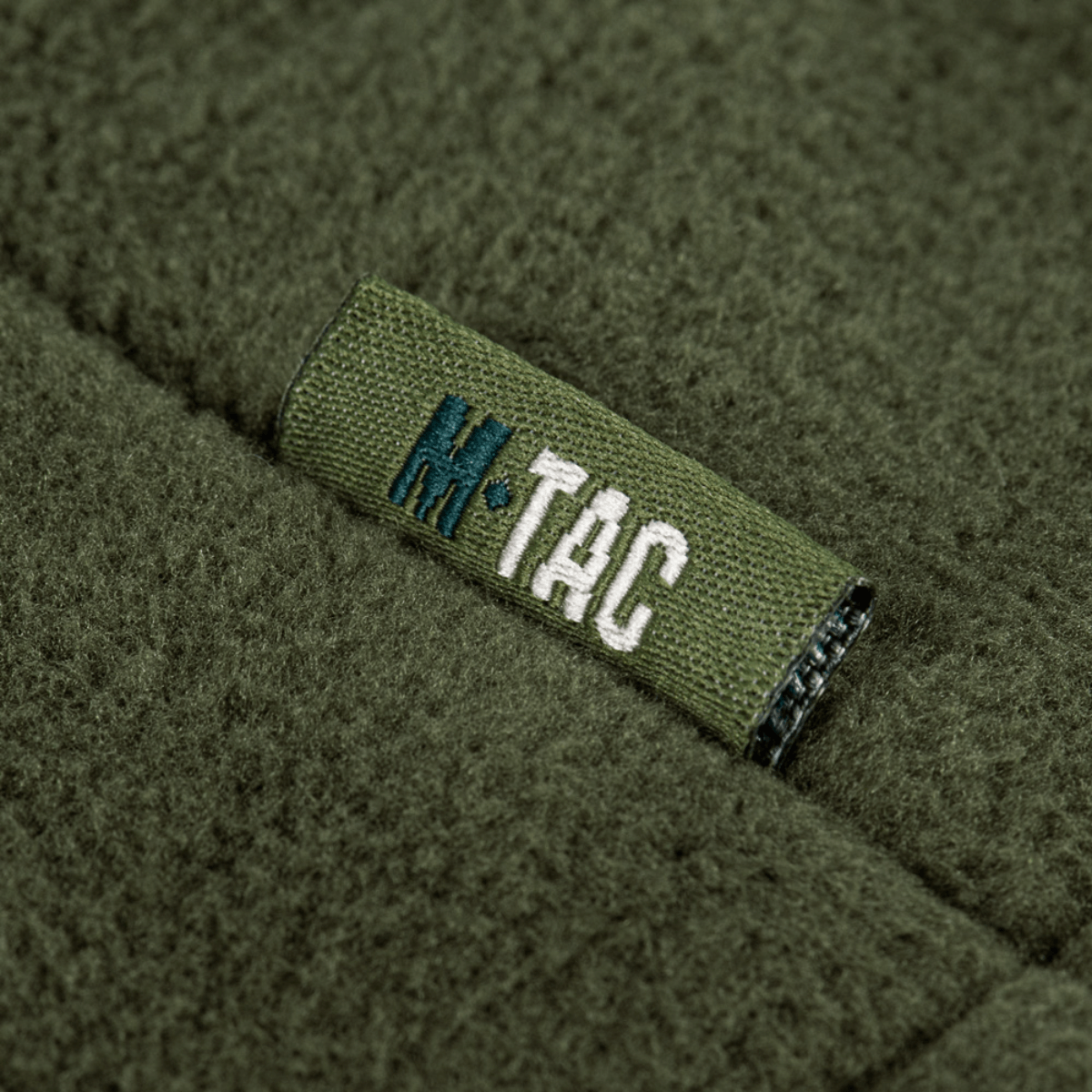 Yksityiskohta M-Tac Combat Fleece Polartec -takista merkin logolla oliivinvihreänä, näyttää muotoilun yksityiskohdat.
