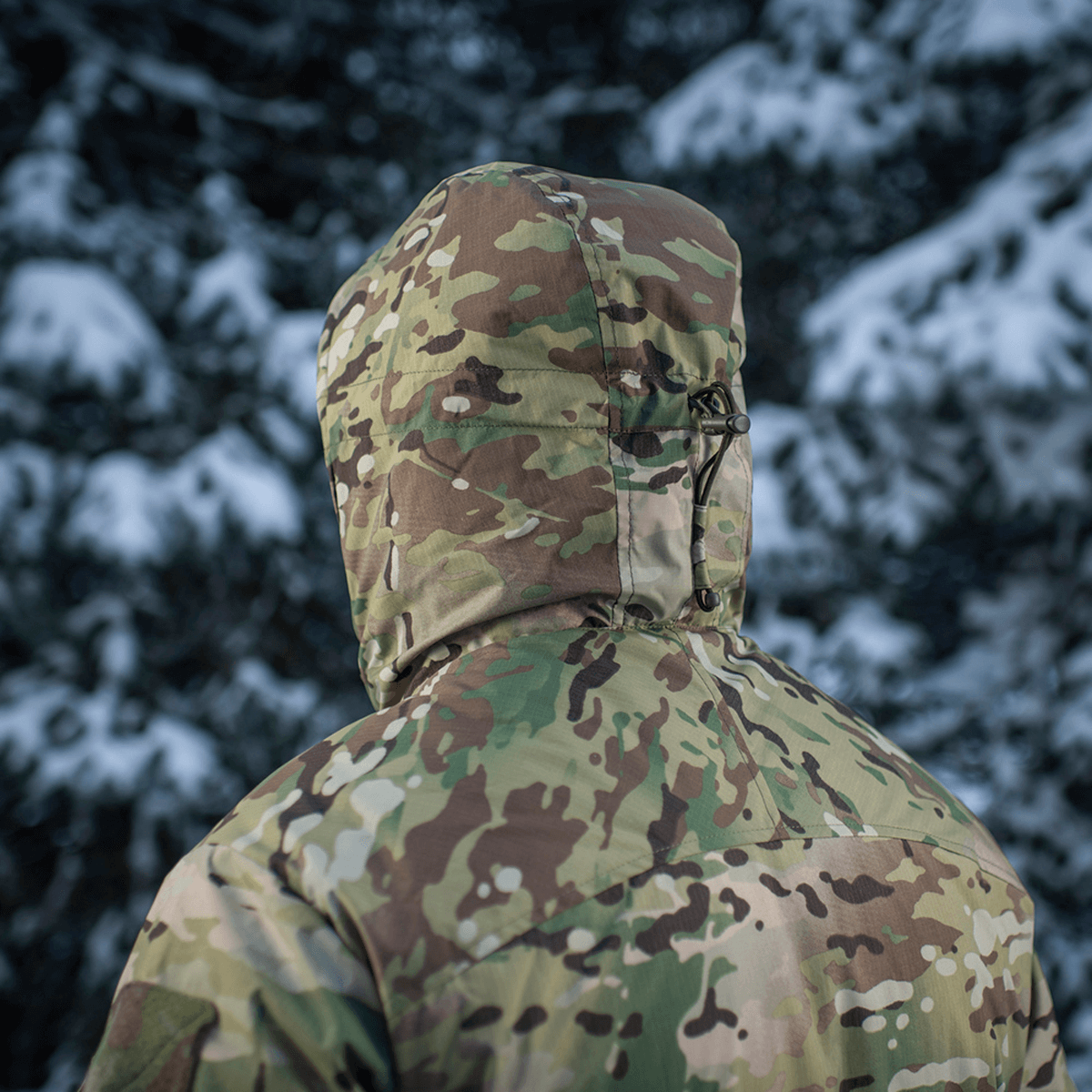 M-Tac Alpha Gen.IV Pro Primaloft -talvitakki Multicam-kankaalla lumisten puiden edustalla, sopii kylmiin sääolosuhteisiin.