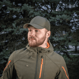 M-Tac Mesh Flex Ripstop Baseball Cap vihreänä, henkilö käyttää sitä armeijaa muistuttavassa takissa metsäalueella.