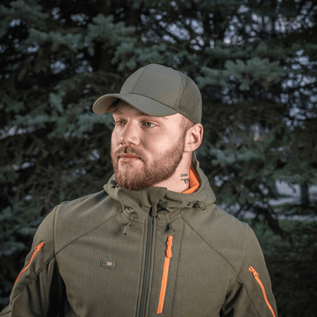 M-Tac Mesh Flex Ripstop Baseball Cap vihreänä, henkilö käyttää sitä armeijaa muistuttavassa takissa metsäalueella.