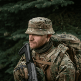 Mies pukeutuneena M-Tac Panama Hat Gen.II NYCO camo-designiin, sopii ulkoiluun ja sotilastyyliin, repun kanssa metsässä.