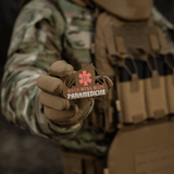 M-Tac Patch Viking Evac sotilaan kädessä, näyttää 'Odin will wait paramedicine' -aiheen. Maastotakki ja taktinen liivi taustalla.
