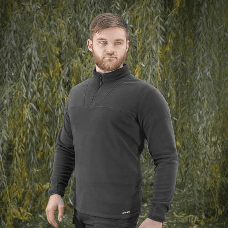M-Tac Delta Polartec fleece-takki mustana puolivetoketjulla miehellä vihreiden pensaiden edessä.