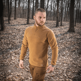 Mies pukeutuneena M-Tac Delta Polartec fleece-takkiin metsässä, seppiavärinen fleece, puolivetoketju, kestävä rakenne, hengittävä muotoilu.