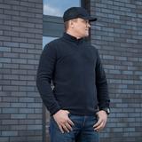 Mies käyttää mustaa M-Tac Delta Polartec -fleece-takkia Polartec®-fleecestä muuriseinän edessä.