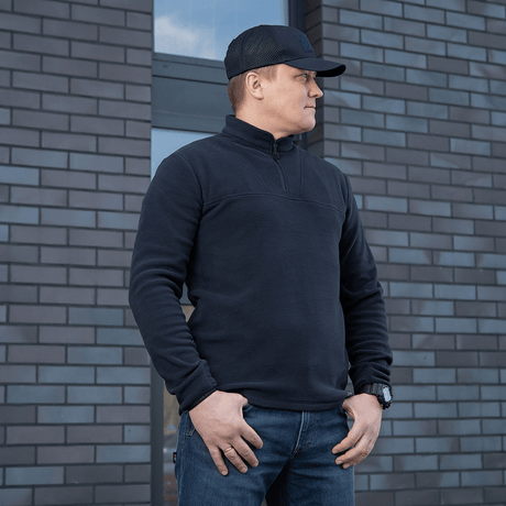 Mies käyttää mustaa M-Tac Delta Polartec -fleece-takkia Polartec®-fleecestä muuriseinän edessä.