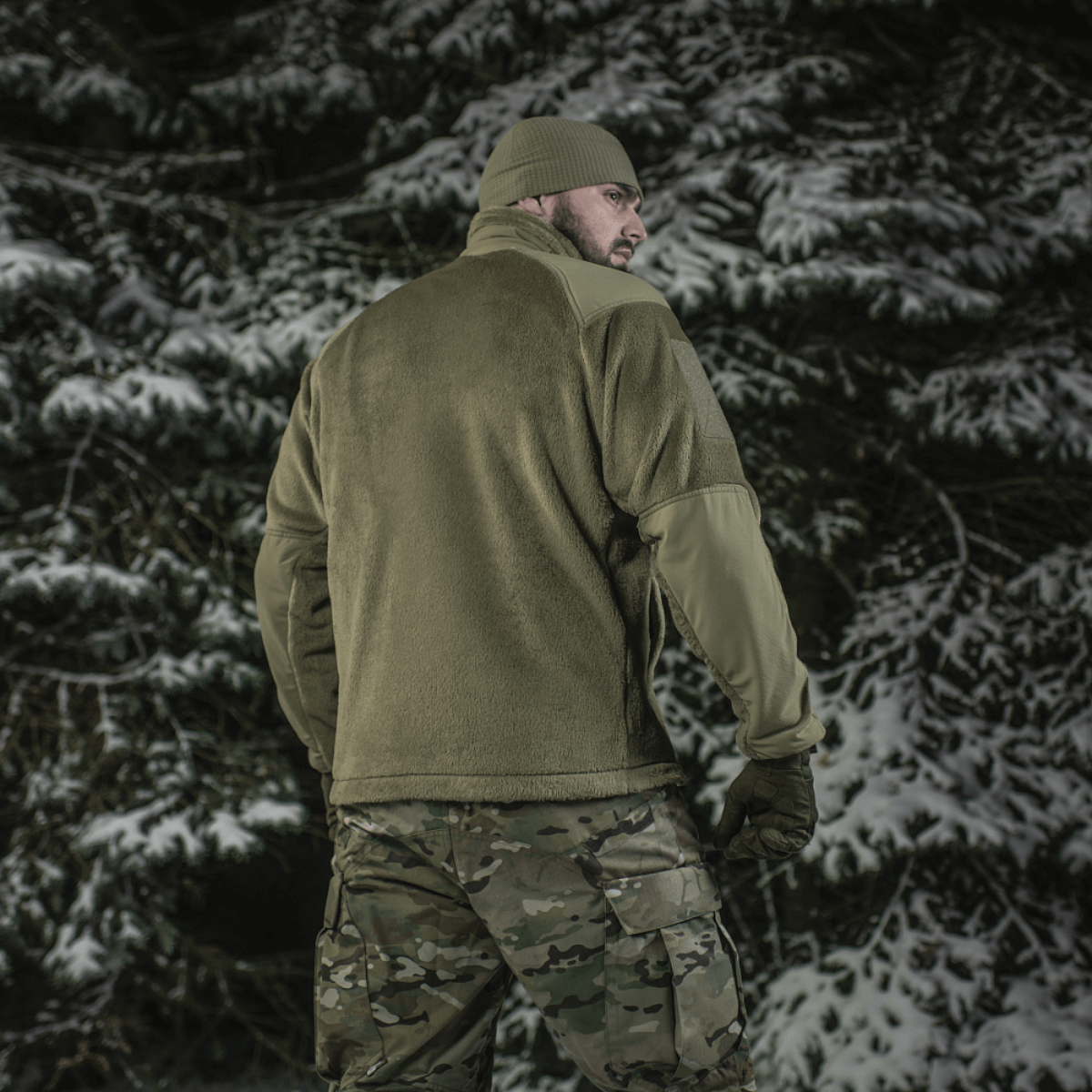 Mies pukeutuneena M-Tac Battle Fleece Polartec -takkiin lumisessa metsässä, nähtynä takaa anatomisella istuvuudella ja maastohousuilla.