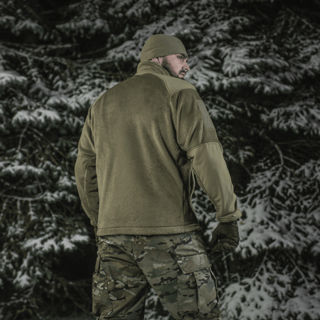 Mies pukeutuneena M-Tac Battle Fleece Polartec -takkiin lumisessa metsässä, nähtynä takaa anatomisella istuvuudella ja maastohousuilla.