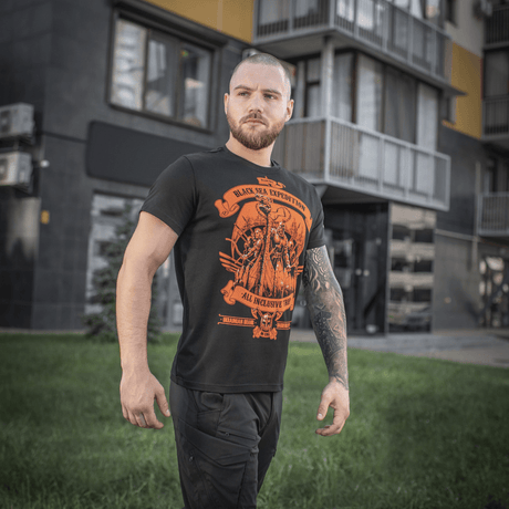 Mies pukeutuneena mustaan M-Tac T-shirt Black Sea Expedition -paitaan kaupunkiympäristössä, näyttää urheilullisen leikkauksen ja uniikin printin.