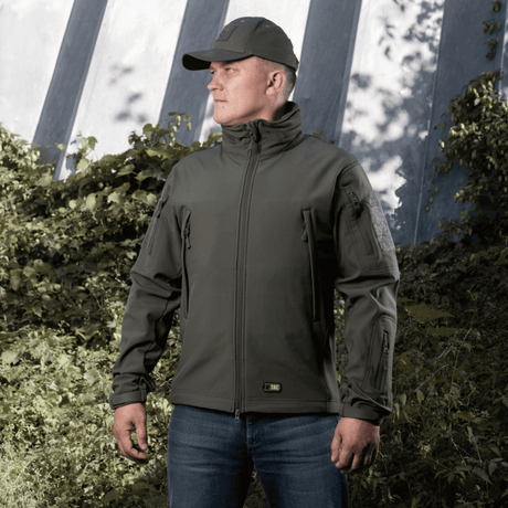 Mies pukeutuneena M-Tac Soft Shell Jakkeen, jossa fleecevuori ja sääsuoja, seisoo ulkona vihreää taustaa vasten.