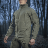 Mies pukeutuneena M-Tac Soft Shell Jakkeen metsäympäristössä, suunniteltu suojaamaan huonolta säältä polyuretaanikalvon ja fleece‑kerroksen avulla.