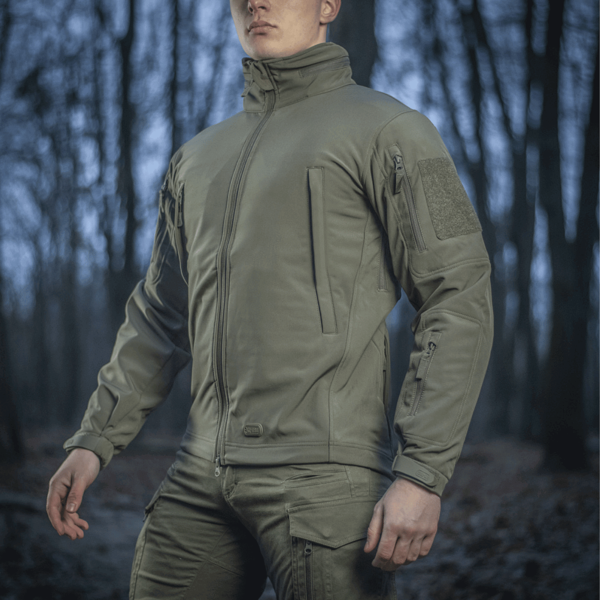 Mies pukeutuneena M-Tac Soft Shell Jakkeen metsäympäristössä, suunniteltu suojaamaan huonolta säältä polyuretaanikalvon ja fleece‑kerroksen avulla.