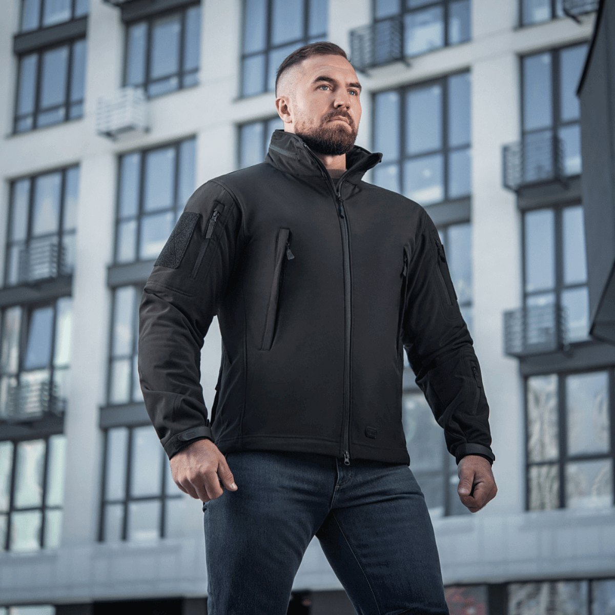 Mies pukeutuneena M-Tac Soft Shell -takkiin seisoo modernin rakennuksen edessä, näyttää takin sääsuojaus- ja eristysominaisuuksia.