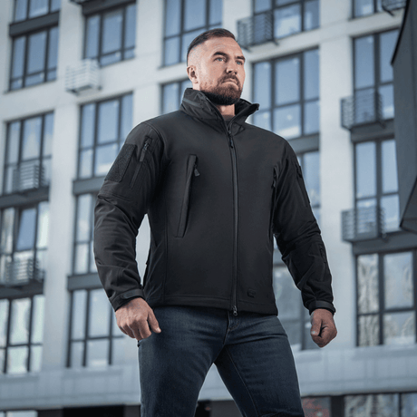 Mies pukeutuneena M-Tac Soft Shell -takkiin seisoo modernin rakennuksen edessä, näyttää takin sääsuojaus- ja eristysominaisuuksia.