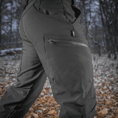 M-Tac Soft Shell Vinter -housut metsässä, mustat, fleece-vuori ja korkea vyötärö, sopivat talviaktiviteetteihin, suojaavat kylmältä ja kosteudelta.