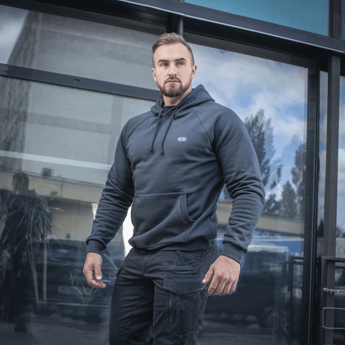 Mies M-Tac Cotton Raglan -hupparissa lasirakennuksen edessä, rennon ja mukavan näköinen, sopii arkeen.