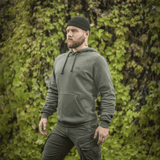 Mies vihreässä M-Tac Cotton Raglan -hupparissa ulkona lehtipeitteisen taustan edessä, rento ja mukava tyyli.