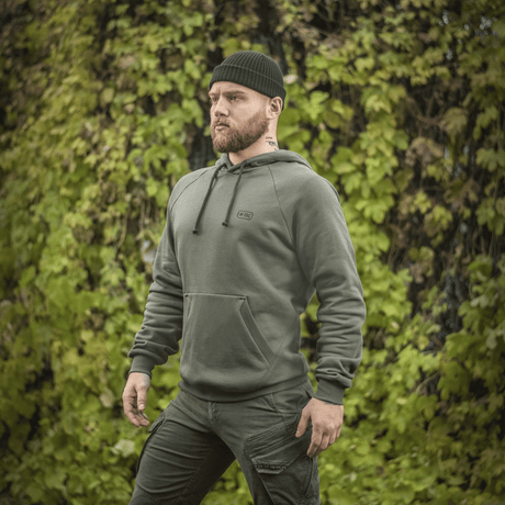Mies vihreässä M-Tac Cotton Raglan -hupparissa ulkona lehtipeitteisen taustan edessä, rento ja mukava tyyli.