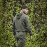 Mies M-Tac Cotton Raglan -hupparissa vihreiden lehtien edessä, näyttää raglanhihat ja hupun, mukava vaatetus.