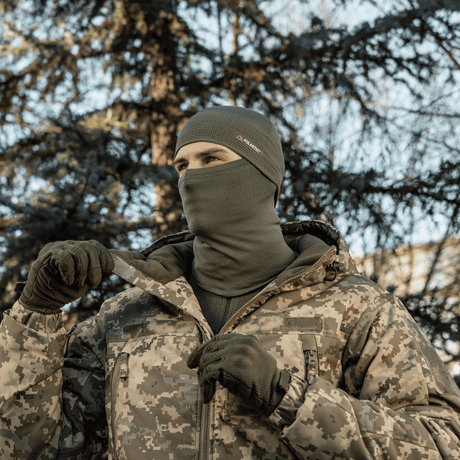 Mies pukeutuneena M-Tac Tube Scarf Polartec Power Grid -huiviin maastokuvioisissa vaatteissa, puiden ympäröimänä, suojaa kylmältä.