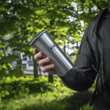 Henkilö pitää M-Tac 450 ml termosmukia venttiilillä vihreässä metsässä